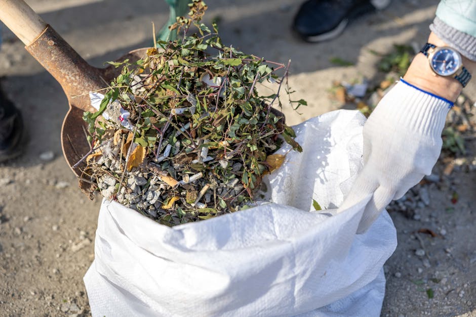 A person wearing a grey sweater, white glove, and a blue watch is seen holding a large white gardening sack filled with clippings, weeds, and small branches. In their left hand, they grip the sack whi